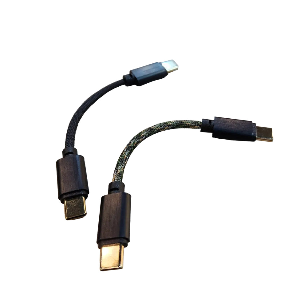 A USB-C to USB-C adapter cable with two connectors, one featuring a black braided cord and the other with a green-and-black patterned cord, both with dark wood-textured housings, displayed on a light wood grain surface.
